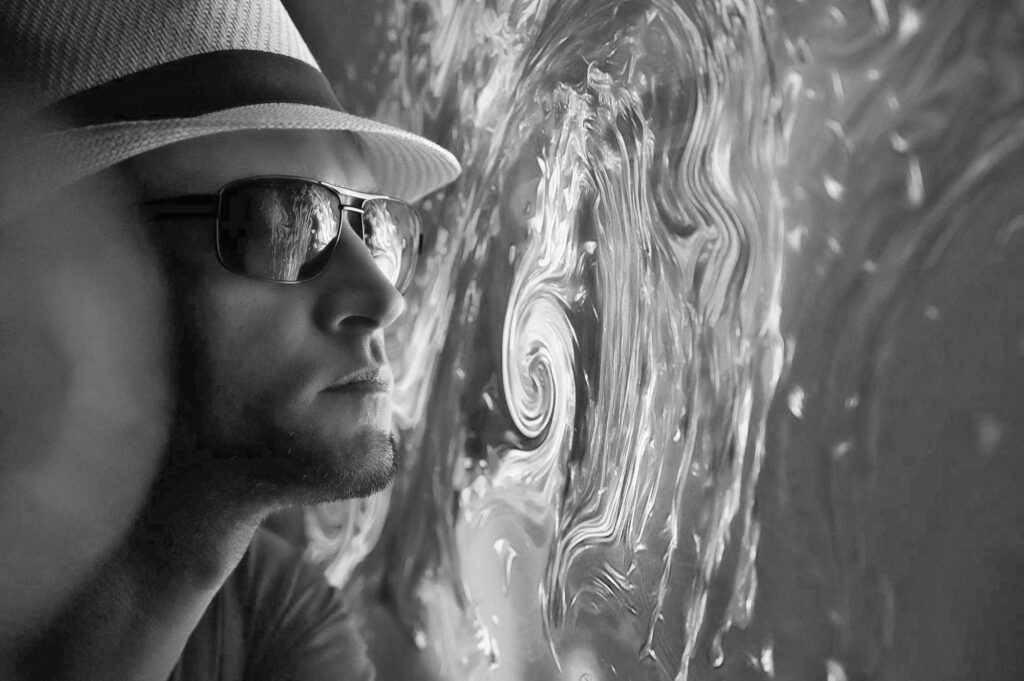 A black-and-white image of a person wearing a hat and sunglasses, gazing at a swirling, abstract pattern reflected in their lenses. The dramatic contrast highlights the interplay between individual focus and complex external influences, symbolizing introspection and the evolving nature of work identity.